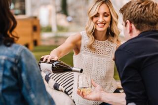 A woman pours wine into a glass, smiling, while sitting with friends outdoors in a relaxed setting, enjoying the moment together.