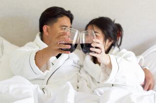 A couple in cozy bathrobes cheers with glasses of red wine while snuggled in a bed, creating an intimate and relaxed atmosphere.