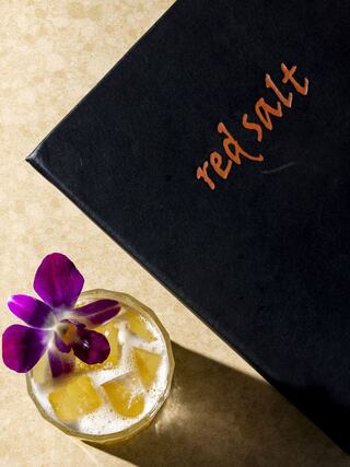 A tropical cocktail garnished with a purple flower sits beside a sleek black menu labeled "red salt" on a light countertop.