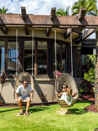 A couple enjoys swinging in stylish chairs outside a modern house, surrounded by lush greenery and a sunny sky.