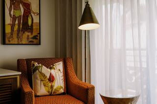 A cozy corner features a patterned armchair with a decorative pillow, a sleek lamp, and sheer curtains letting in soft light.