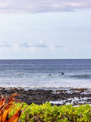Surfers catch waves in a calm ocean, framed by rocks and lush greenery under a cloudy sky, creating a serene coastal scene.