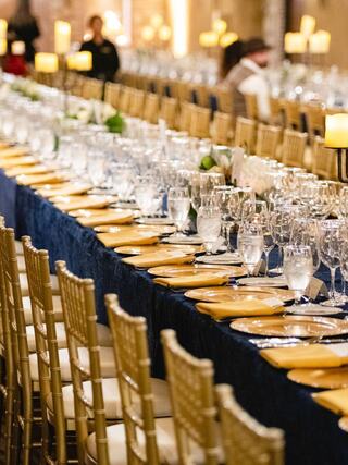 A grand dining setup features elegantly arranged tables with golden plates, sparkling glassware, and candlelight, creating an inviting atmosphere.