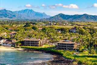 A coastal scene features beachfront resorts amid lush palm trees, with majestic mountains in the background under a bright blue sky.