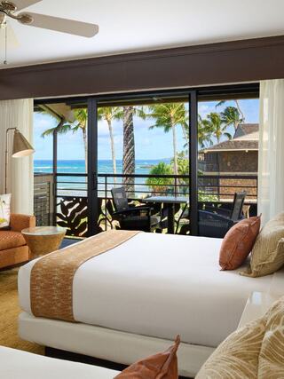 A cozy bedroom features two beds, tropical decor, and a balcony with stunning ocean views and palm trees. Perfect for relaxation.