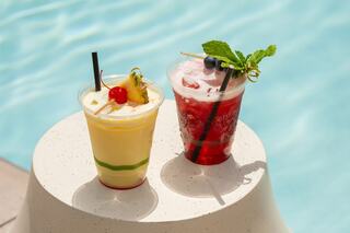 Two colorful drinks sit on a white table by a sparkling pool, one yellow with cherry and pineapple, the other red topped with mint and blueberries.