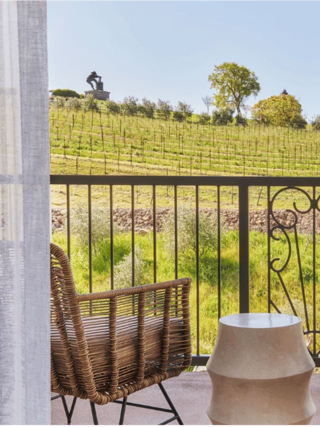 A cozy balcony features a wicker chair and a round table, overlooking lush vineyards and a sculpted figure in the distance under a clear sky.