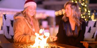 Two smiling friends enjoy roasting marshmallows over a glowing fire pit, surrounded by festive lights on a cozy night.