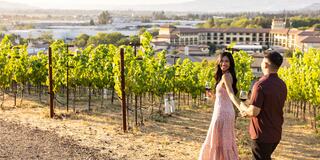 A couple strolls through a vineyard, holding glasses of wine, with a scenic view of a town and mountains in the background during sunset.
