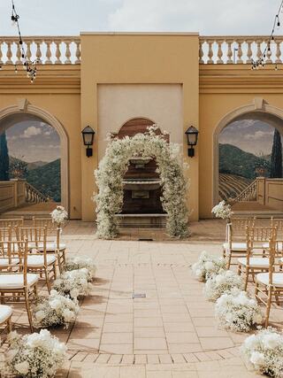 An elegant wedding venue features a floral arch, gold chairs, and decorative arrangements, set against a scenic mural backdrop.