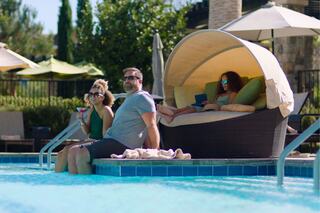 A man and a woman relax by a pool, enjoying drinks, while a girl sits in a cabana, focused on her device, in a sunny setting.