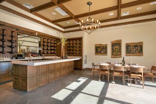 A stylish tasting room featuring a wooden bar, elegant chandeliers, and a cozy dining area with artwork, illuminated by natural light.