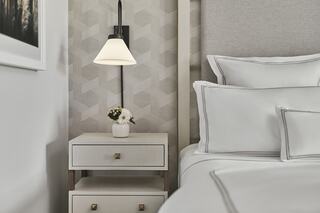A cozy bed features crisp white linens beside a stylish nightstand with a small vase of flowers and a modern wall lamp, set against a patterned wall.