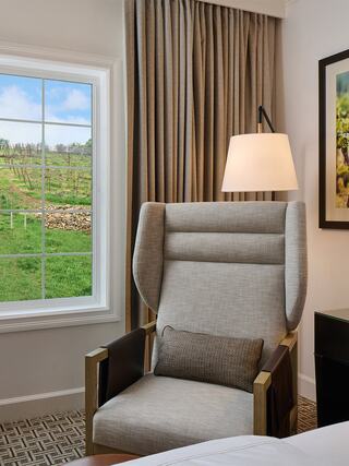 A cozy hotel room features a stylish armchair beside a window with views of a vineyard, complemented by soft curtains and modern decor.