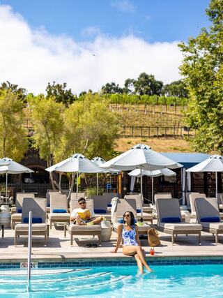 Two people relax by a poolside, surrounded by lounge chairs and shaded umbrellas, with lush vineyards in the background and a clear blue sky.