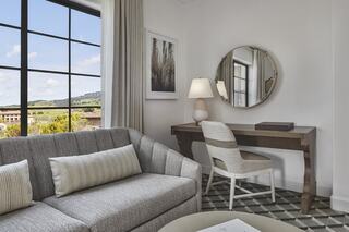 A cozy, modern living space features a gray sofa, a wooden desk with a lamp, and a large window showcasing a scenic view outside.
