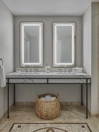Elegant hotel bathroom features double sinks, modern illuminated mirrors, and a stylish woven basket, complemented by neutral tones and marble accents.