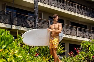 A shirtless man in colorful shorts holds a surfboard, standing among vibrant greenery in front of a stylish building.