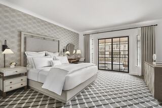 A stylish hotel room features a cozy bed, elegant decor, a patterned rug, and a balcony with draped curtains, creating a welcoming atmosphere.