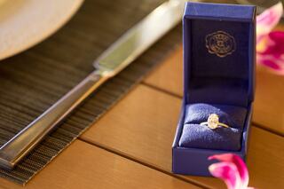 A couple in white robes joyfully pours champagne on a balcony, with a beautiful ocean view in the background. Sunlight enhances the moment.