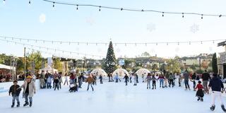 A lively ice skating rink filled with families and friends, adorned with festive lights and a large Christmas tree in the background.