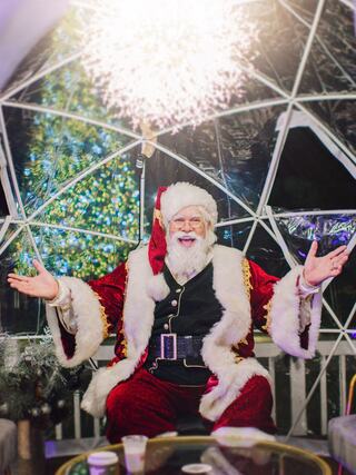 A jolly figure in a Santa costume smiles warmly, surrounded by festive decorations and sparkling lights in a cozy igloo-like setting.