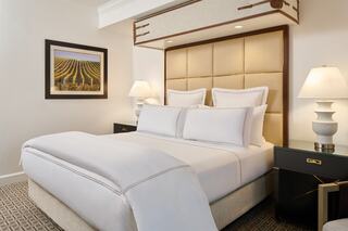 A stylish hotel room features a plush bed with white linens, elegant lamps, and a framed landscape art piece, creating a cozy ambiance.