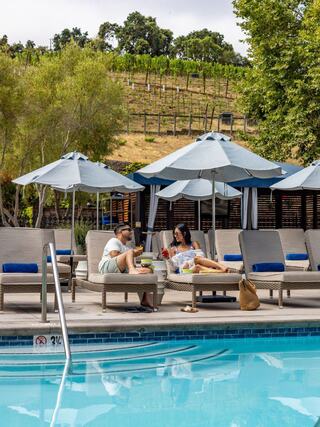A couple relaxes by a poolside, enjoying drinks and snacks under umbrellas, with a scenic vineyard backdrop.