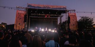 A large crowd enjoys a live performance at an outdoor concert, with colorful banners and stage lights set against a twilight sky.