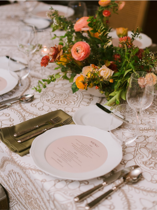 An elegantly set dining table features floral arrangements, fine tableware, and a menu on a decorative tablecloth.