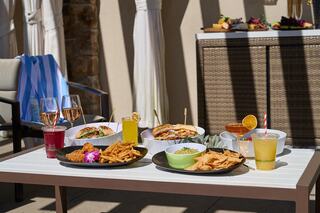 A sunlit table showcases a variety of delicious dishes and drinks, including colorful cocktails, fried foods, and vibrant garnishes.