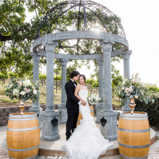 A couple embraces under a decorative gazebo surrounded by vineyards, adorned with floral arrangements and wooden barrels, capturing a romantic moment.