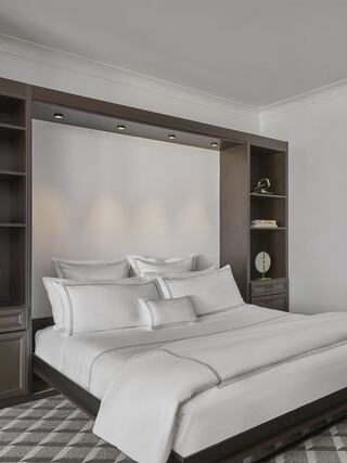 A sleek, modern bedroom features a neatly made bed with white linens, accented by soft lighting and a stylish bookshelf backdrop.