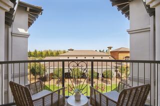 A serene balcony view featuring two wicker chairs, a small table, and a lush courtyard with greenery under a clear blue sky.