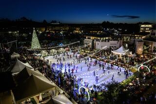 A bustling ice skating rink surrounded by festive lights and crowds, with a tall Christmas tree illuminated against a twilight sky.