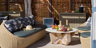 A cozy outdoor lounge area features wicker seating, a marble coffee table with a colorful fruit platter, and a serene, shaded ambiance.