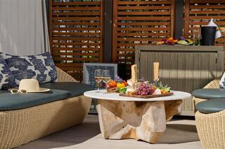 A cozy outdoor seating area featuring natural wood furniture, a centerpiece of fruit, and glasses, surrounded by stylish decor and sunlight.