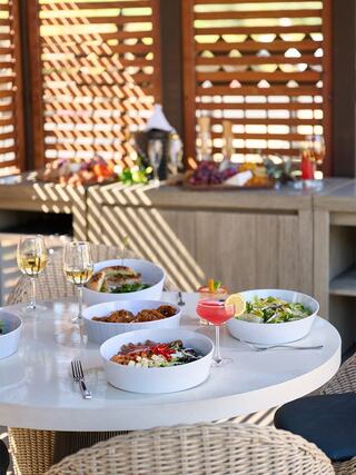 A stylish outdoor dining setup features plates of colorful food, cocktails, and wine, bathed in warm sunlight with a cozy ambiance.