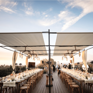 An elegant outdoor dining setup features long tables adorned with candles and flowers, under a canopy, surrounded by scenic views.