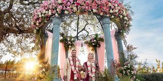 A couple stands beneath a floral arch adorned with pink blooms, celebrating their wedding as the sun sets in a picturesque vineyard setting.