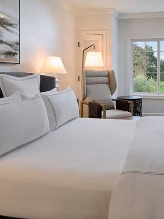 A cozy bedroom featuring a neatly made bed, soft lighting from lamps, a stylish armchair, and a view of greenery outside the window.