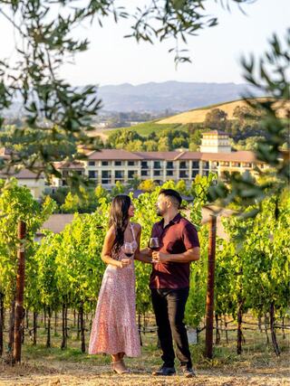 A couple enjoys a moment among lush vineyards, holding wine glasses, with rolling hills and a picturesque resort in the background.