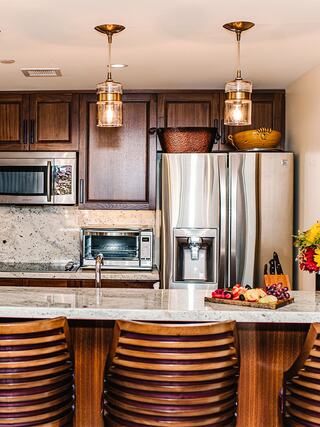 A modern kitchen features wooden cabinets, stainless appliances, and a granite countertop, with stylish pendant lights and a fruit centerpiece.