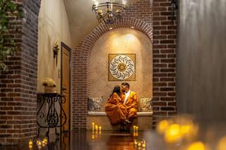 A cozy, romantic setting with a couple in orange robes, sitting close together by glowing candles and a decorative wall.