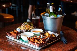 A tray filled with delicious appetizers like wings, nachos, and dips, accompanied by a bucket of assorted beers and a glass of soda.