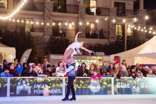 A couple performs an elegant ice skating routine, captivating a lively crowd filled with festive lights and holiday cheer.