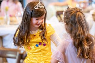 Two young girls in playful outfits, one in a yellow dress with stars and a tiara, share a joyful moment at a festive gathering.
