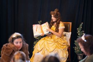 A woman in a yellow gown reads a storybook to children, surrounded by festive decorations and engaged listeners.