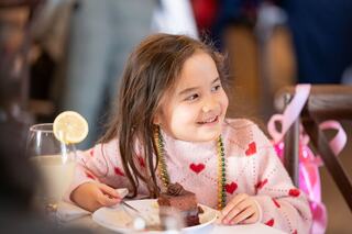 A young girl in a pink sweater with hearts enjoys a slice of cake, smiling brightly. A drink with a lemon slice sits nearby.