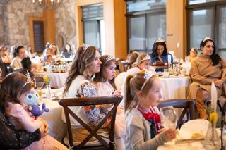 A group of children and adults wearing tiaras seated at elegantly set tables, enjoying a festive gathering in a cozy venue.